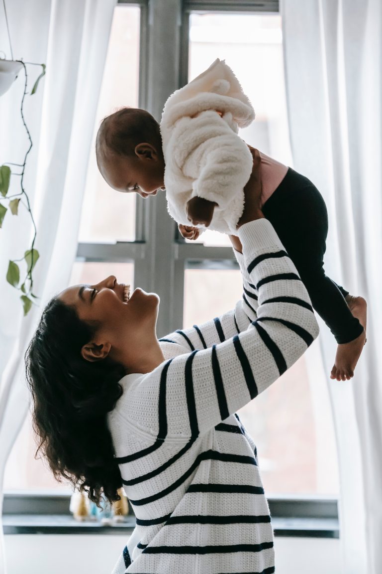 Side view of cheerful mother and cute black baby in hands looking at each other while standing near window
